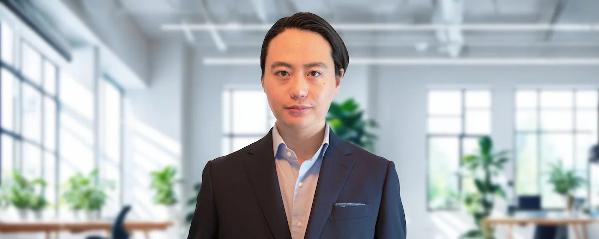明るいオフィスを背景に、スーツを着たアジア人男性がカメラをまっすぐ見つめているポートレート写真です。ビジネスシーンに適したフォーマルな印象を与えます。