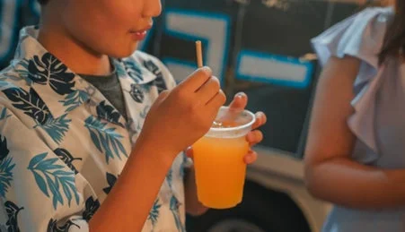 夏らしい柄のシャツを着た子供が、ストローを使ってオレンジ色の飲み物を飲んでいるクローズアップ。爽やかな夏の雰囲気が感じられる。