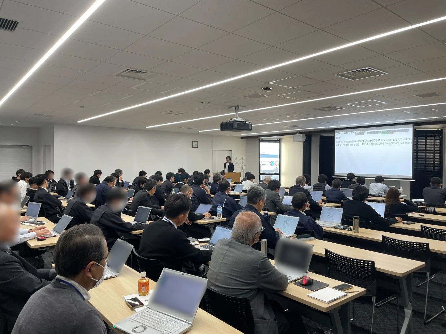 多くのビジネスパーソンがノートパソコンを使い、プロジェクター画面を見ながらセミナーに参加している様子。