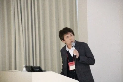 マイクを手に白いポディウムの前に立つ男性が、聴衆に向かって話している様子です。スーツを着用しており、講演会やセミナーでのプレゼンテーションの場面と考えられます。