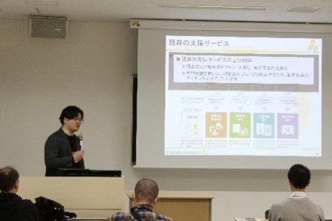 会議室で男性がプロジェクターを使ってプレゼンテーションを行っています。スライドには「既存の支援サービス」と題され、インフラ整備計画や技術開発・運用支援に関する内容、SDGsのような目標を示すアイコンが表示されています。