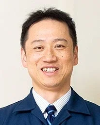 青いスーツとストライプのネクタイを着用した、笑顔の中年男性の顔のアップです。明るい背景の前で撮影されており、真面目さと親しみやすさを兼ね備えた印象を与えます。