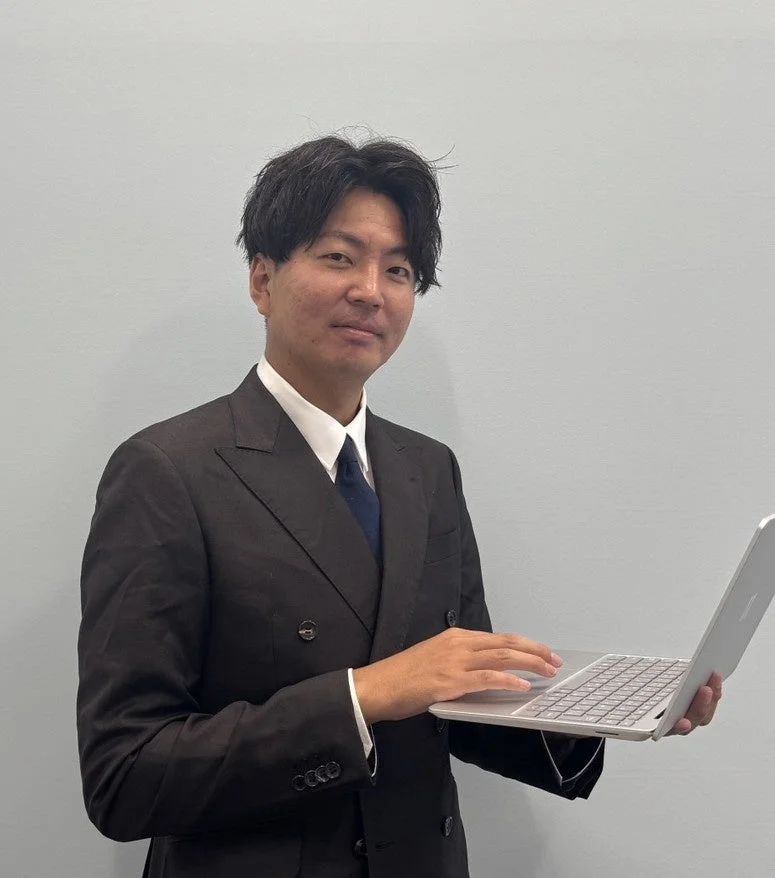 ダークスーツを着た男性がノートパソコンを手に立ち、穏やかな表情でカメラを見ている