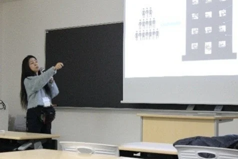 教室で、一人の女性がプロジェクターに映し出された図(人型のアイコンと建物)を指しながらプレゼンテーションを行っています。