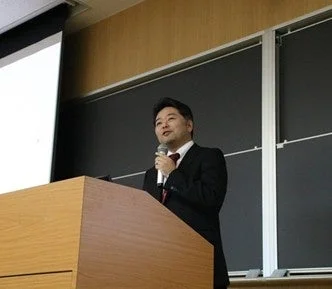 スーツを着た男性が、木製の演台に立ち、マイクを手に聴衆に向かって話している様子。背景にはプロジェクタースクリーンと黒板が見え、大学の講義室やセミナー会場のような場所で講演を行っている場面です。