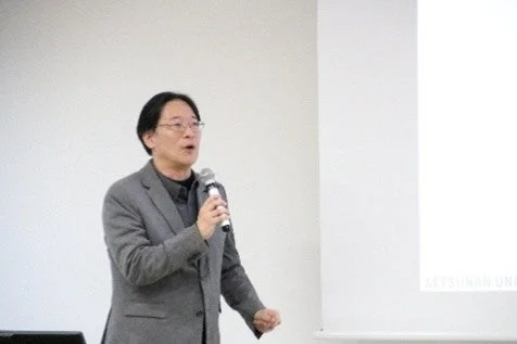 スーツを着た男性がマイクを片手に、白いスクリーンを背景に講演を行っている様子。真剣な表情で聴衆に語りかけている。