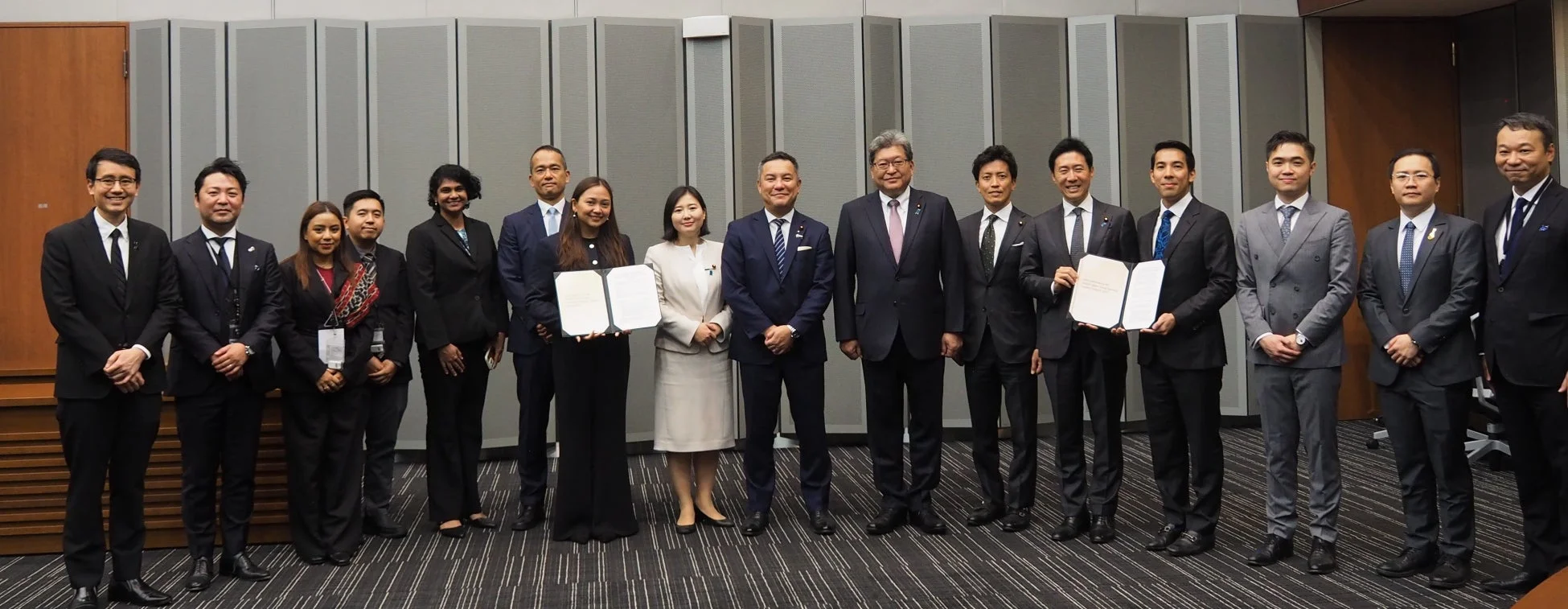 ビジネススーツを着た複数の男女が整列し、一部が書類を手にしている集合写真