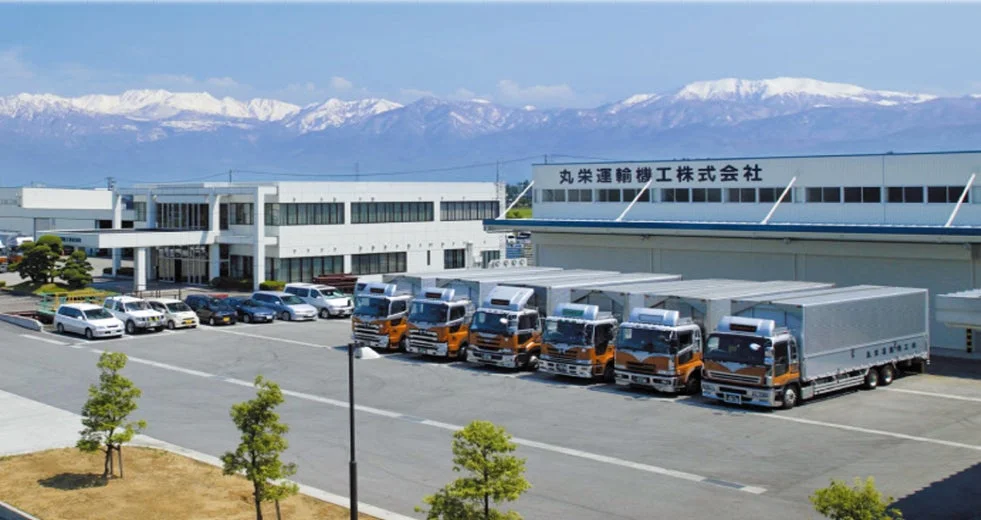 雪山を背景に広がる丸栄運輸機工株式会社の施設と駐車場です。数台の配送トラックと乗用車が停められており、広々とした敷地には建物が複数見えます。晴れた日の風景です。