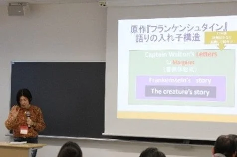 女性がフランケンシュタインの物語構造についてプレゼンテーションをしています。スクリーンには「語りの入れ子構造」としてウォルトン船長の手紙、フランケンシュタイン、クリーチャーの物語が階層的に示され、聴衆が耳を傾けています。