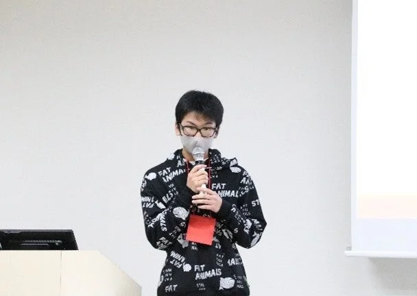 マスクと眼鏡をかけた男性が、マイクを持って発表している様子です。白い壁とスクリーンを背景に、セミナーか講演会のような場所でプレゼンテーションを行っています。