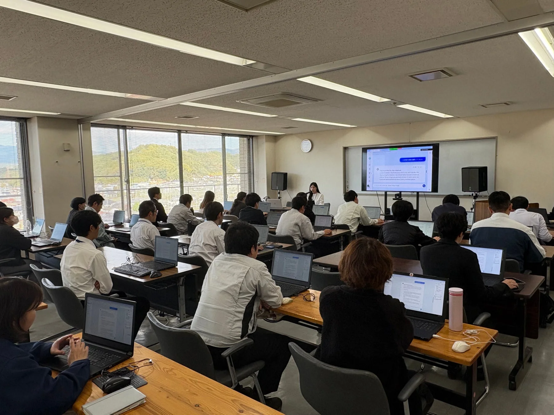 教室で多くの受講生がノートパソコンを使用しながら講義や研修を受けている様子。前方には講師が立ち、スクリーンには「何か質問してください」と表示されている。