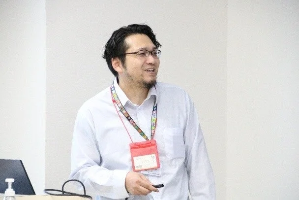 眼鏡と髭を蓄えた男性が白いシャツを着用し、IDカードを首から下げて話している様子です。屋内での会議やプレゼンテーションの場面と見られ、笑顔で何かを説明しているようです。