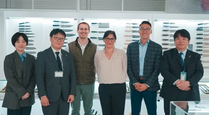 男女6人が笑顔で並び、背景には商品が展示されたショールームのような空間が広がる集合写真です。ビジネスシーンでのチームや訪問者の様子を捉えています。