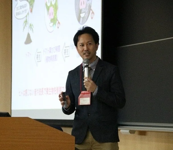 スーツ姿の男性がマイクとリモコンを持ってプレゼンテーションを行っています。背後のスクリーンには、イラストと日本語のテキストが表示されており、何らかの研究や技術に関する発表のようです。