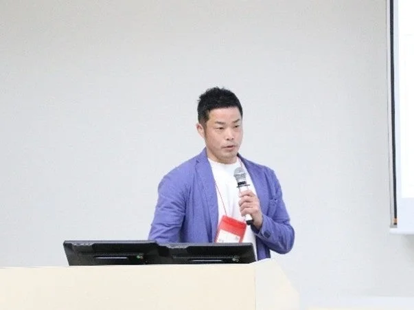 青いジャケットを着た男性が、マイクを持って壇上でプレゼンテーションまたは講演を行っています。会議室やセミナー会場のような場所で、熱心に話している様子がうかがえます。