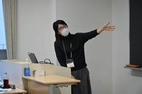 マスクを着用した女性が講演台の前に立ち、右手を伸ばして何かを指差している。会議室のような場所でプレゼンテーションを行っている場面と見られる。