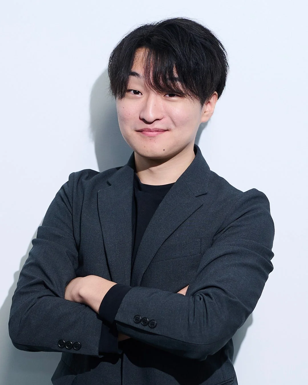 白い背景の前で、ダークグレーのスーツジャケットを着用し、腕を組んで微笑む若いアジア人男性のポートレート