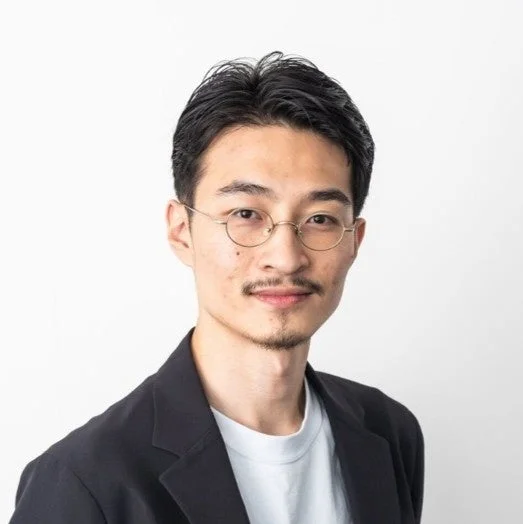 白背景の前で、黒いジャケットと白いTシャツを着用し、丸い眼鏡と髭を蓄えたアジア人男性の上半身ポートレート。カメラをまっすぐ見つめています。