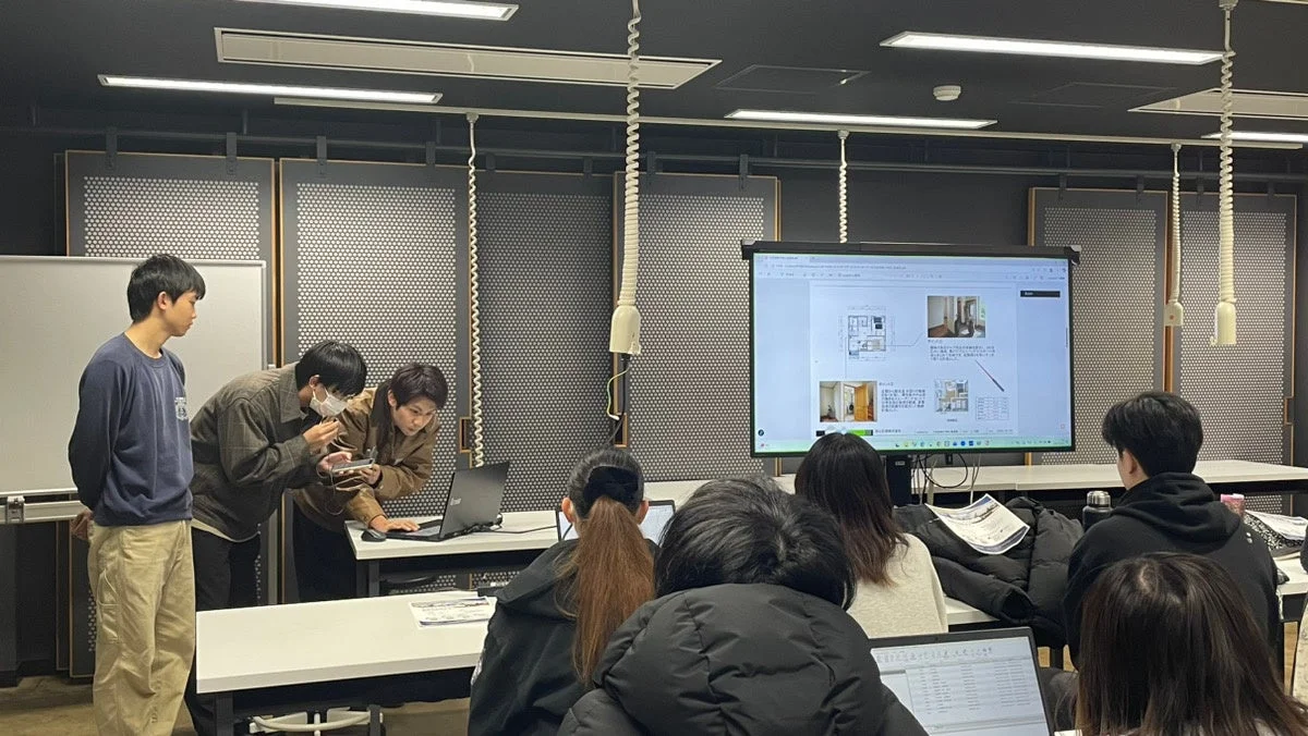 教室で学生たちが集まり、大きなモニターに表示された間取り図やインテリアの写真を見ながら、学習や議論を行っている様子