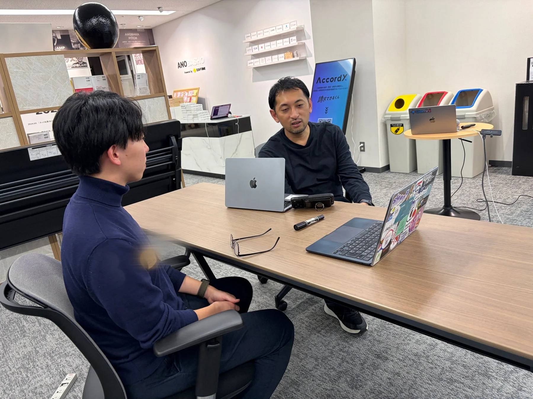 オフィス環境で2人の男性がノートパソコンを前に打ち合わせをしている様子。背景には「ANOBASHO」や「AccordX」などのビジネス関連の表示が見られ、活発な議論が交わされているようです。