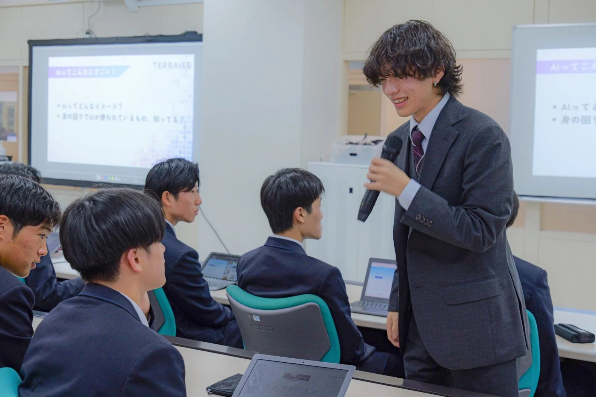 スーツを着た若い男性が教室でプレゼンテーションを行っており、マイクを片手に笑顔で話しています