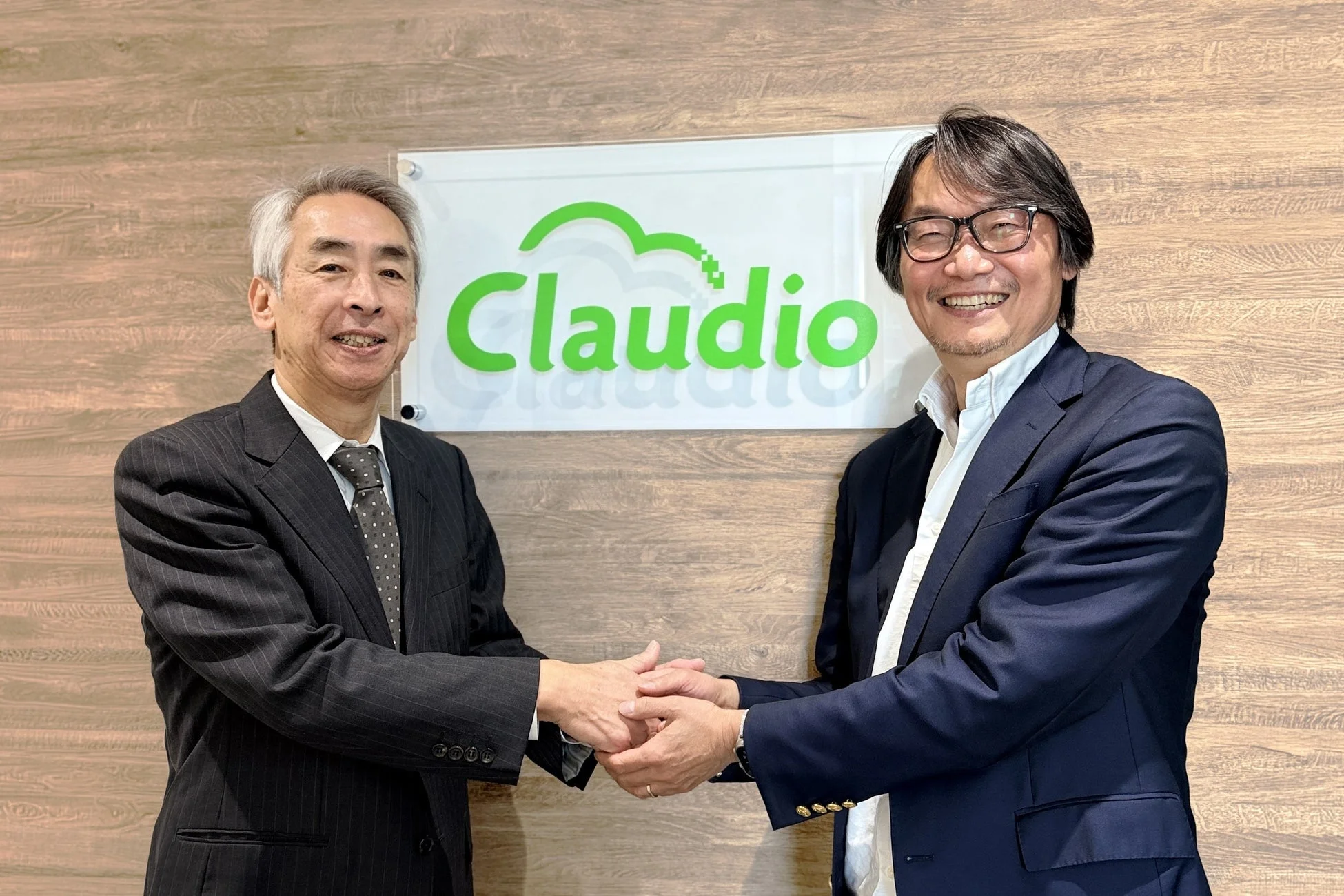 ビジネススーツを着た2人の男性が笑顔で握手をしている。背景には「Claudio」のロゴがあり、ビジネス上の合意や協力関係を示唆するプロフェッショナルな雰囲気の画像である。