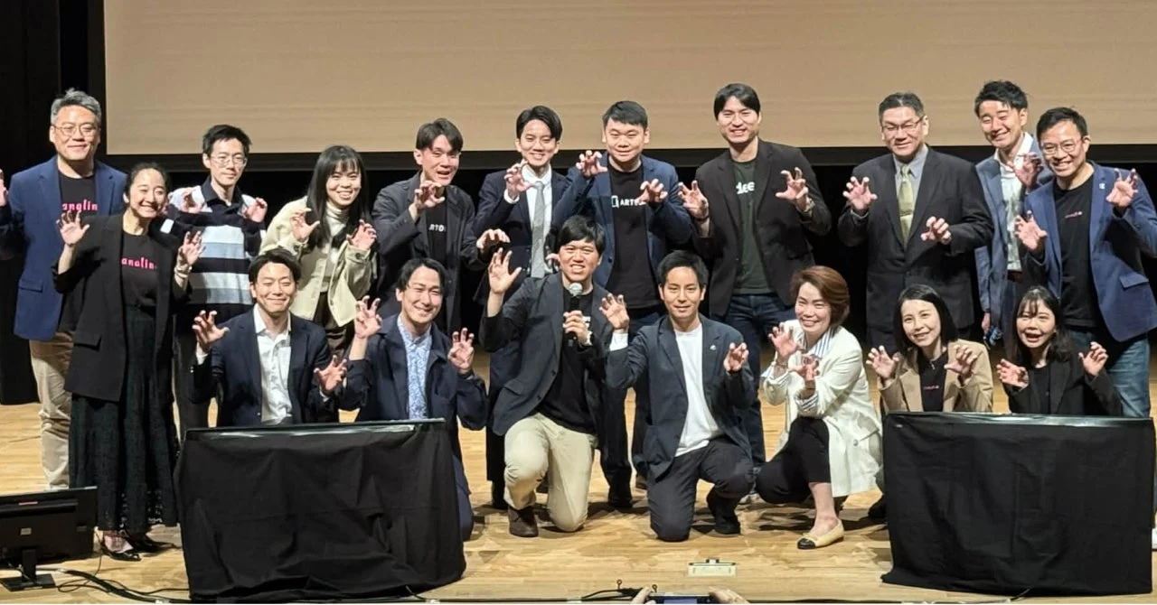 ステージ上で多くの人々が「猫の手」のようなポーズをとって笑顔で写る集合写真