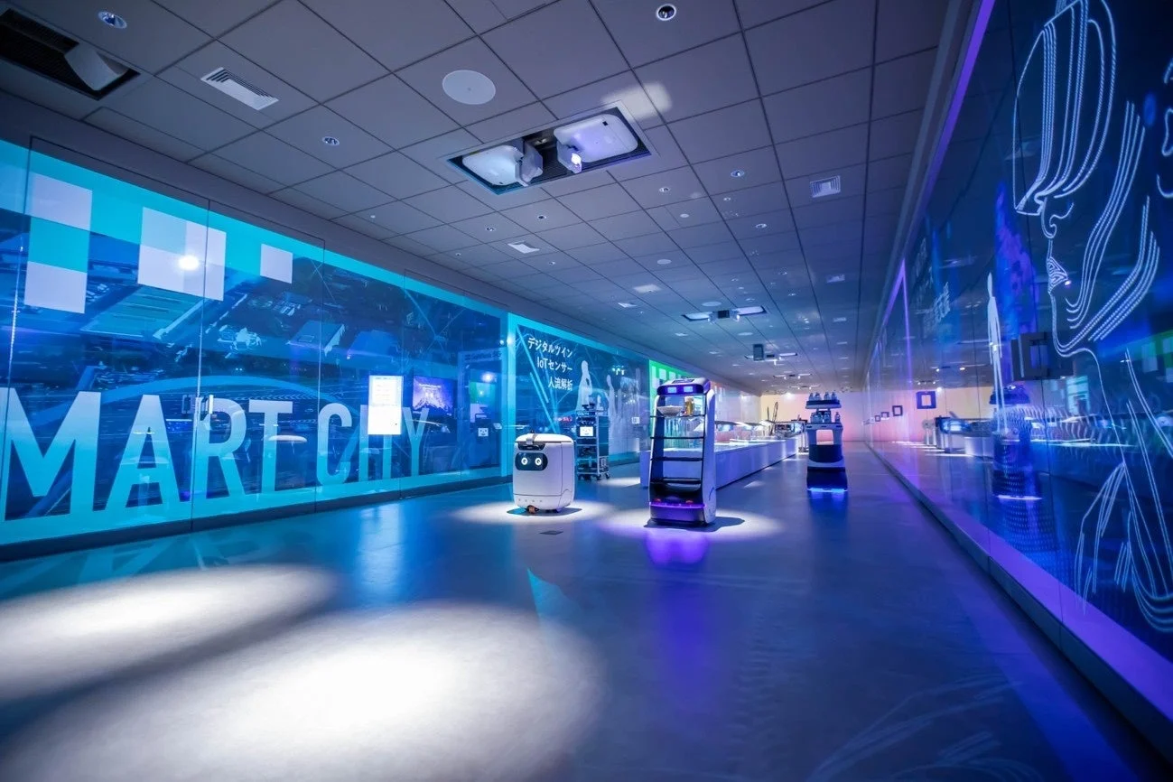 スマートシティのコンセプトを示す未来的な展示ホールで、ロボットが複数設置され、壁面にはデジタルツインやIoTセンサー、人流解析に関する情報が映し出されている。青と紫の照明が特徴的な空間。
