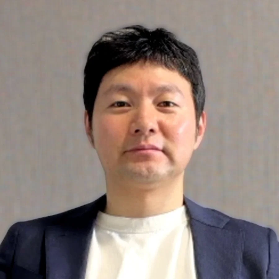 紺色のジャケットと白いTシャツを着用したアジア人男性のポートレート。彼はカメラをまっすぐ見ており、表情は真顔で、背景はシンプルなグレーの壁です。