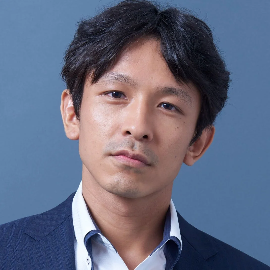 青い背景を背に、ネイビーのスーツジャケットと白いシャツを着用した男性のバストアップポートレートです。カメラを見つめる真剣な表情が印象的です。