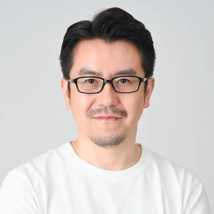 白いTシャツを着て眼鏡をかけた中年のアジア人男性のポートレートです。薄いひげを蓄え、カメラに向かって穏やかな表情をしています。