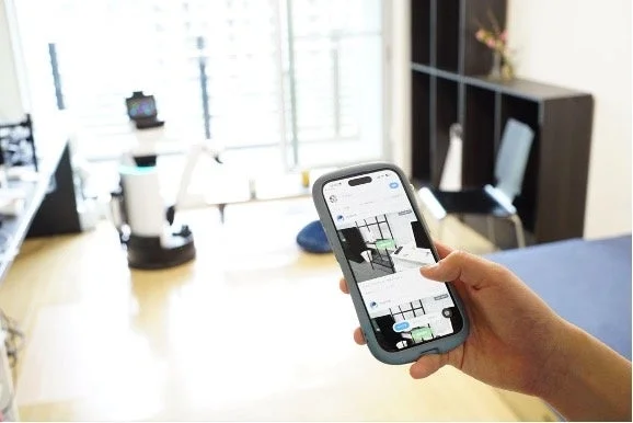 スマートフォンでロボットのアプリを操作する手元と、奥に立つロボット