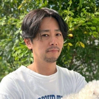 緑豊かな背景の屋外で、白いTシャツを着た髭の男性が、白いふわふわした犬を抱えているポートレート