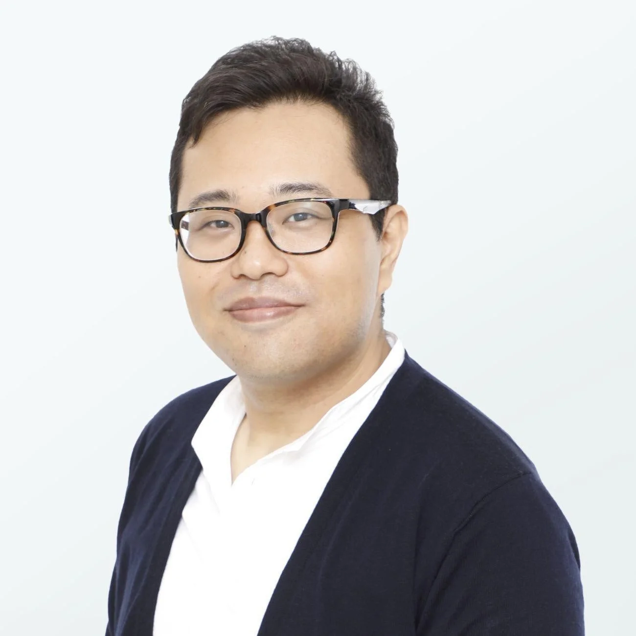 眼鏡をかけた男性が、白いシャツの上に紺色のカーディガンを羽織り、カメラに向かって穏やかに微笑んでいる上半身のポートレート画像