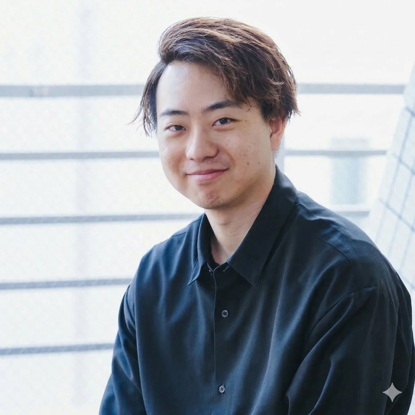 黒いシャツを着た若い男性が、明るい室内でカメラに向かって優しく微笑んでいます。自然光が差し込む窓を背景に、リラックスした雰囲気のポートレートです。