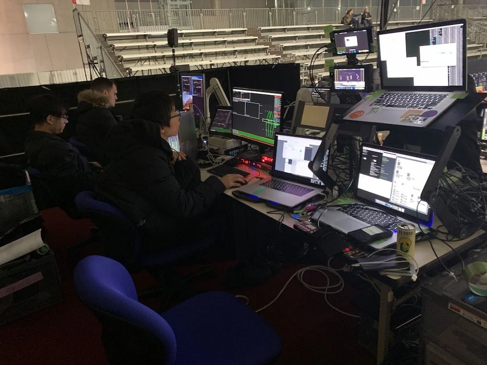 イベント会場のオペレーションルームで、複数のスタッフが多数のモニターとパソコンを操作している様子。複雑な機材が並び、専門的な作業が行われていることが伺えます。