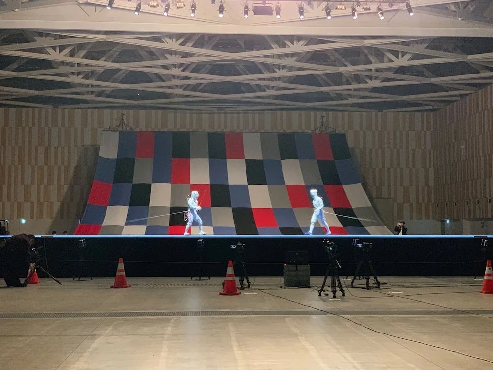 広いアリーナのステージで、フェンシング選手らしき2人がパフォーマンス中。背景はカラフルなチェッカー柄で、ステージ前には複数の撮影用カメラが並び、イベントの様子を捉えている。