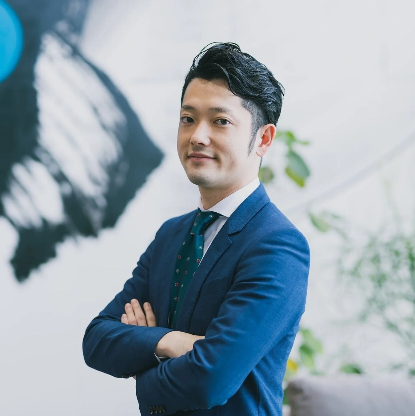 青いスーツを着用したアジア人男性が腕を組み、穏やかな表情でカメラを見ています。背景には抽象的なアートと植物が見えるオフィスのような空間が広がっており、プロフェッショナルな雰囲気を醸し出しています。