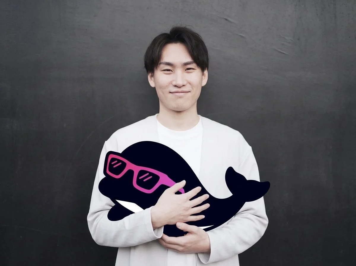 笑顔の若い男性が、ピンクのサングラスをかけた黒いクジラのぬいぐるみを抱えている。シンプルな黒い背景の前で、彼は白いTシャツとカーディガンを着用している。