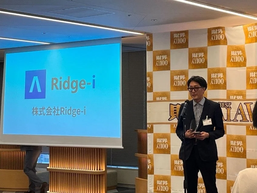 受賞式の様子、マイクを持つ男性と「Ridge-i 財界 AI100 BEST AWARD」のロゴ