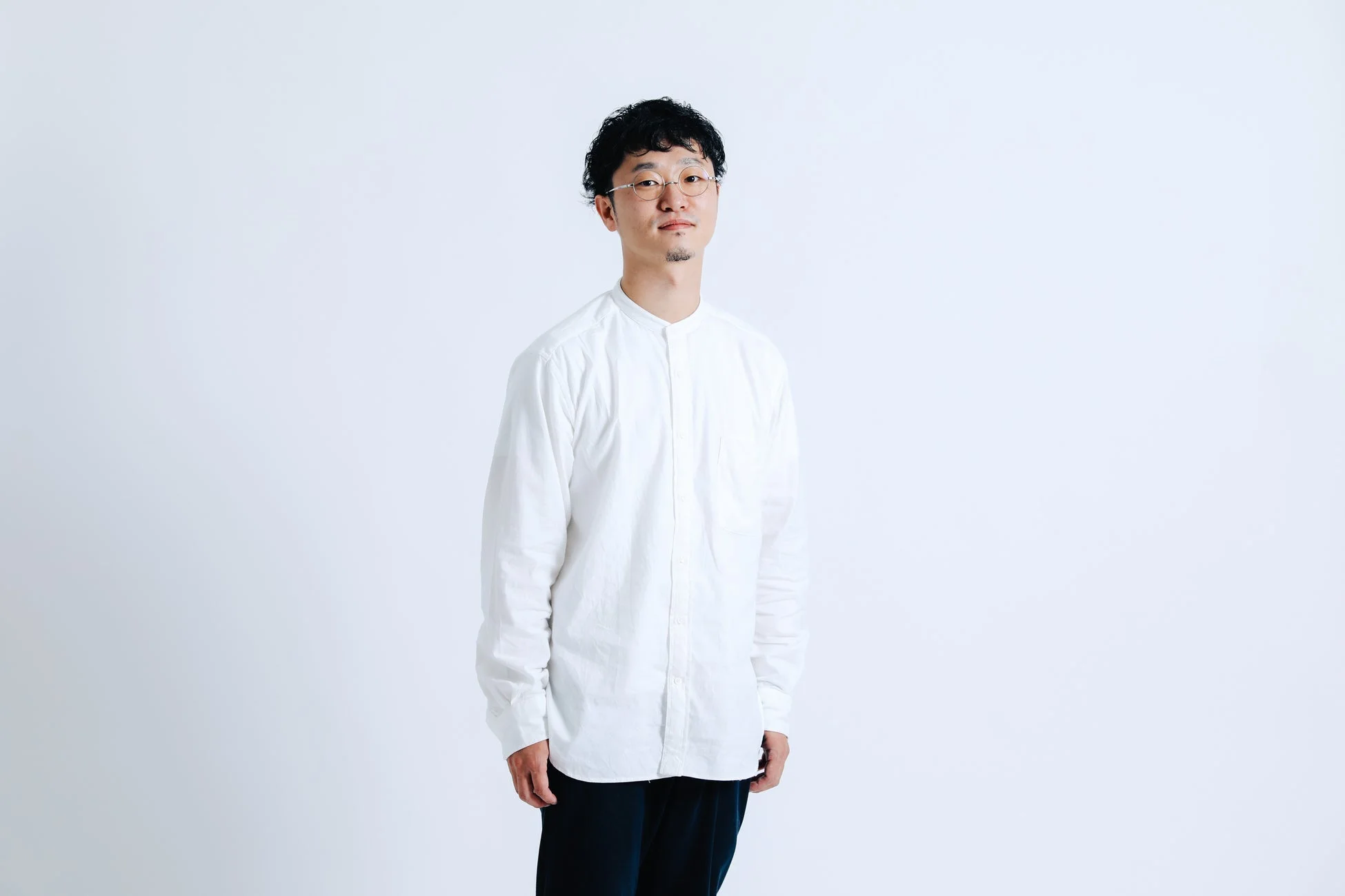 白い背景の前に立つ、白いシャツと黒いパンツを着用したアジア人男性のポートレート。メガネをかけ、短い巻き毛で、カメラに向かって穏やかに微笑んでいます。