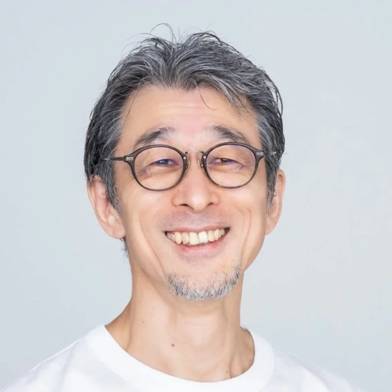 白いTシャツを着て眼鏡をかけた中高年の日本人男性のポートレート写真です。白髪と顎髭が特徴的で、明るい背景の前で親しみやすい笑顔を見せています。