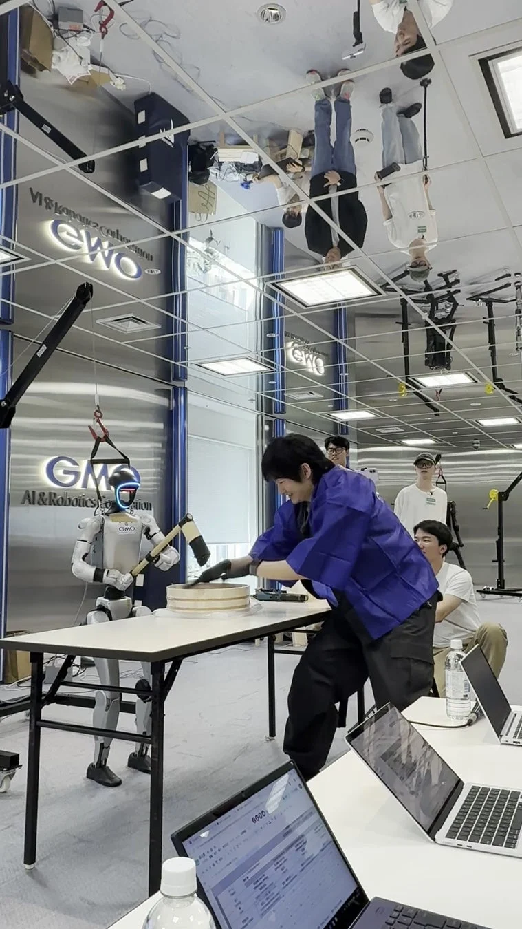 現代的なオフィス環境で、人間とロボットが協力して餅つきを行う様子が捉えられています。