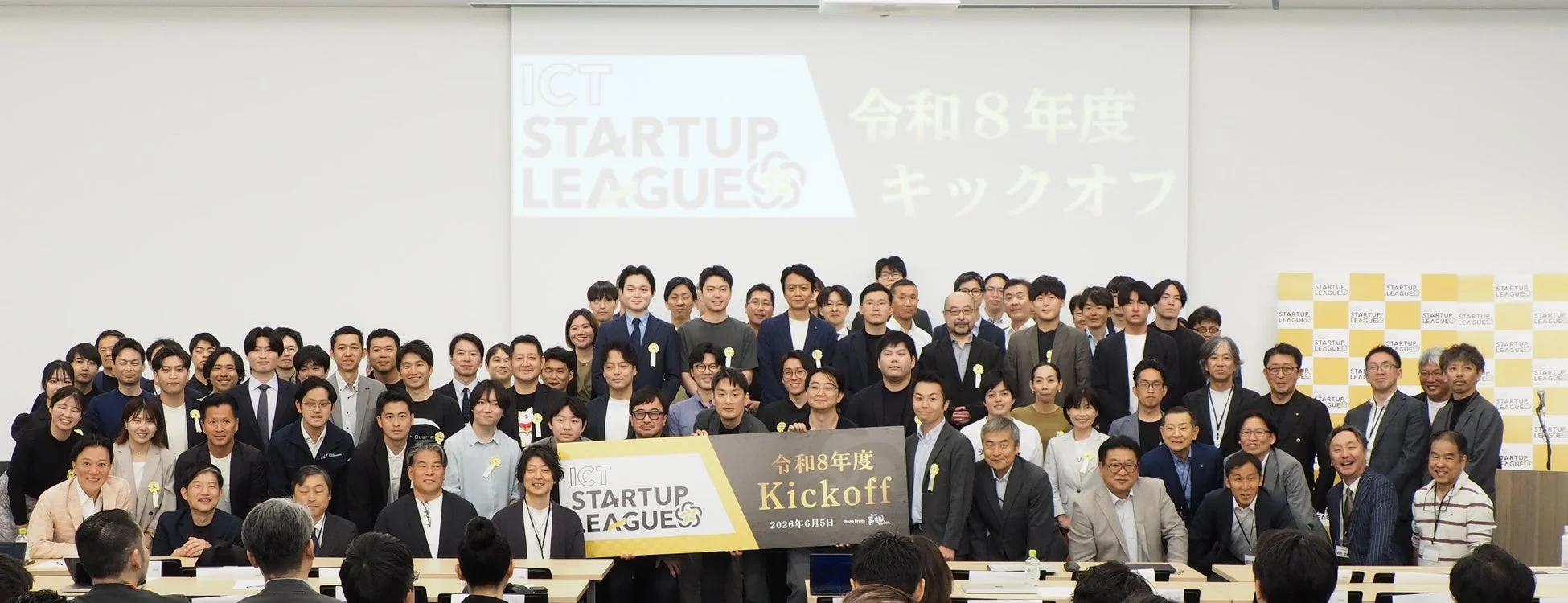 「ICT STARTUP LEAGUE 令和8年度 キックオフ」と書かれたスクリーンとバナーの前で、多数の参加者が集合写真を撮っている様子
