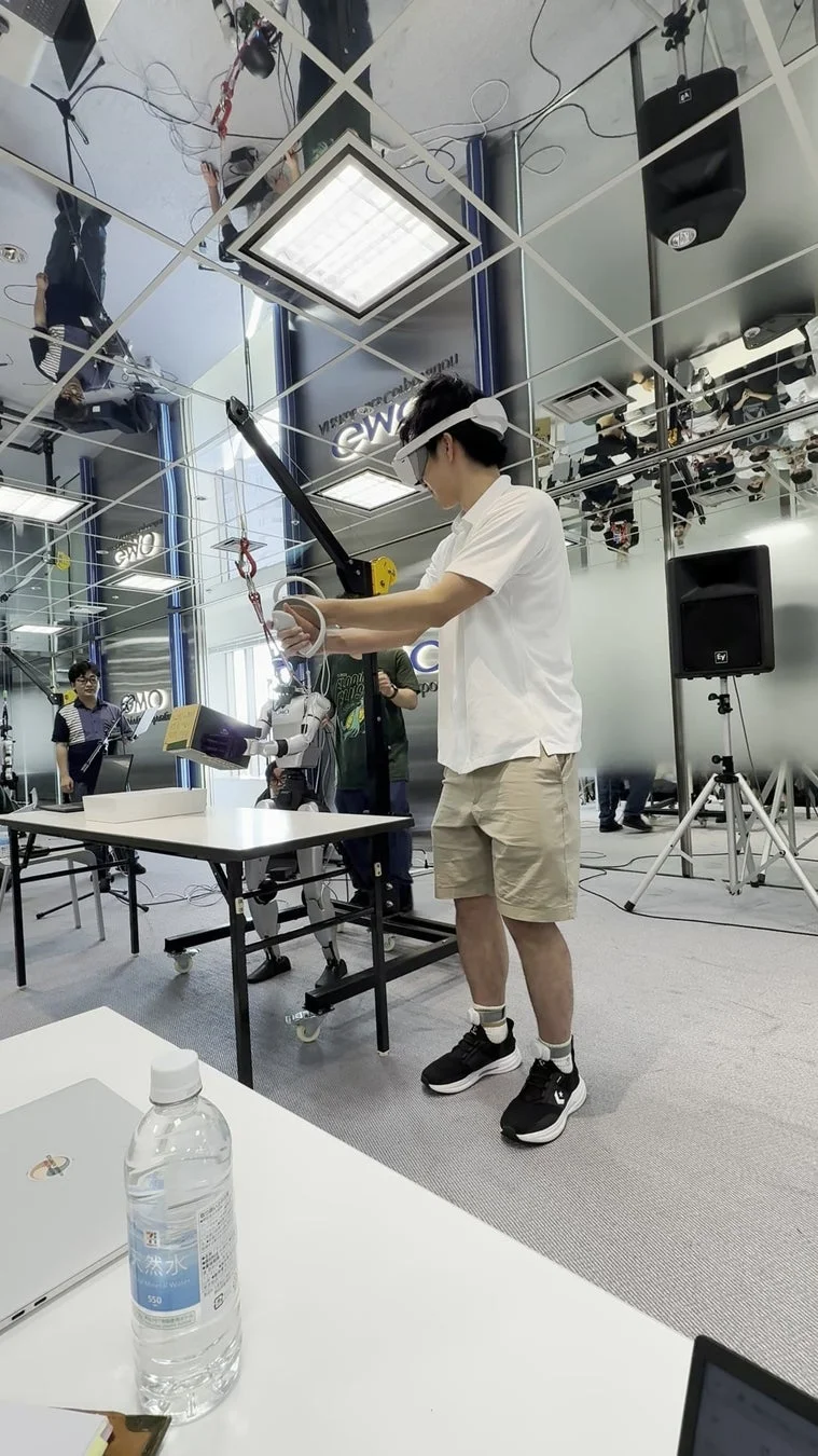 VRヘッドセットを装着した人物が、ミラー張りの天井が特徴的な空間でヒューマノイドロボットを操作しています。
