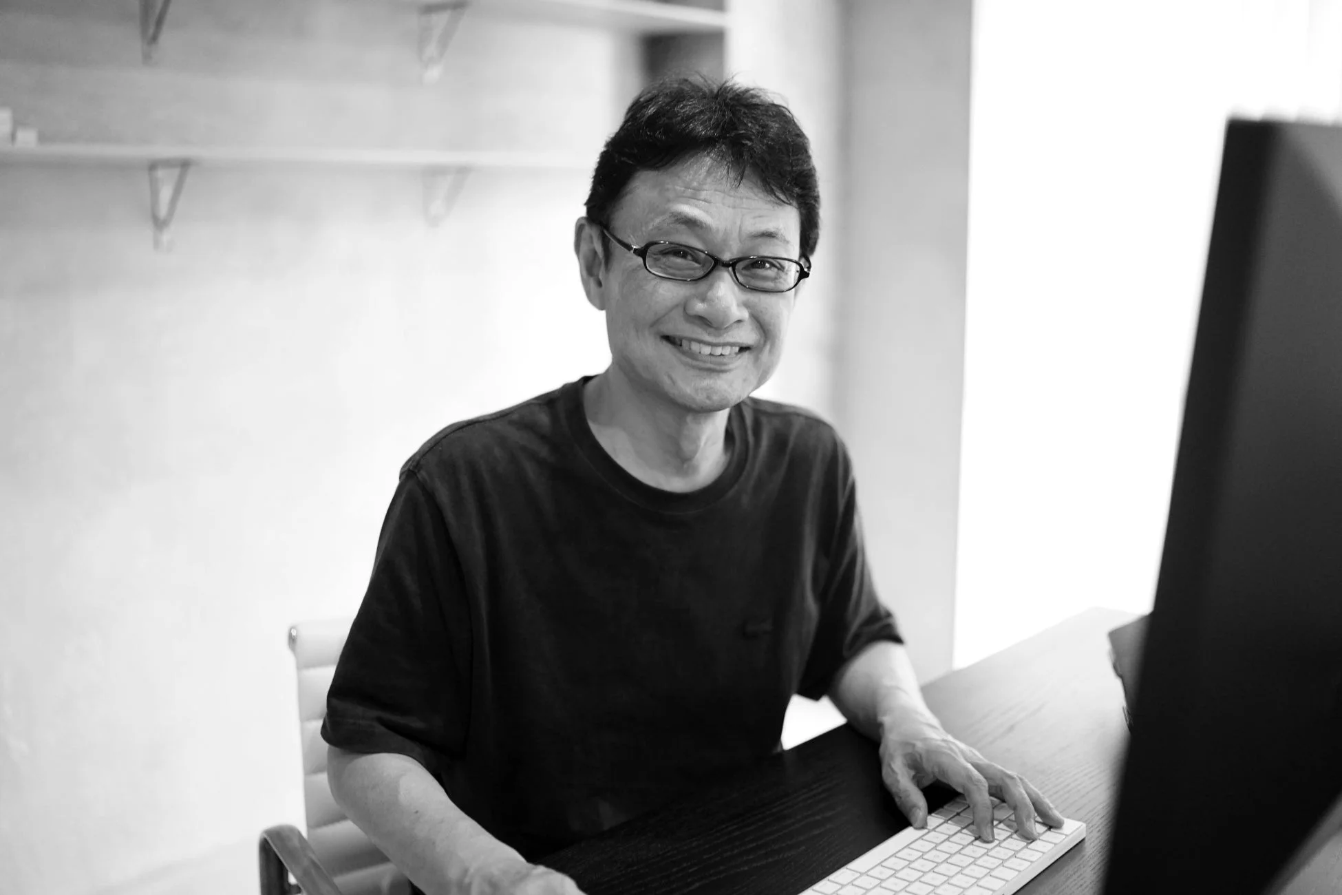 眼鏡をかけたアジア系の男性が、デスクに座り、キーボードに手を置きながら笑顔でカメラを見ているモノクロ写真