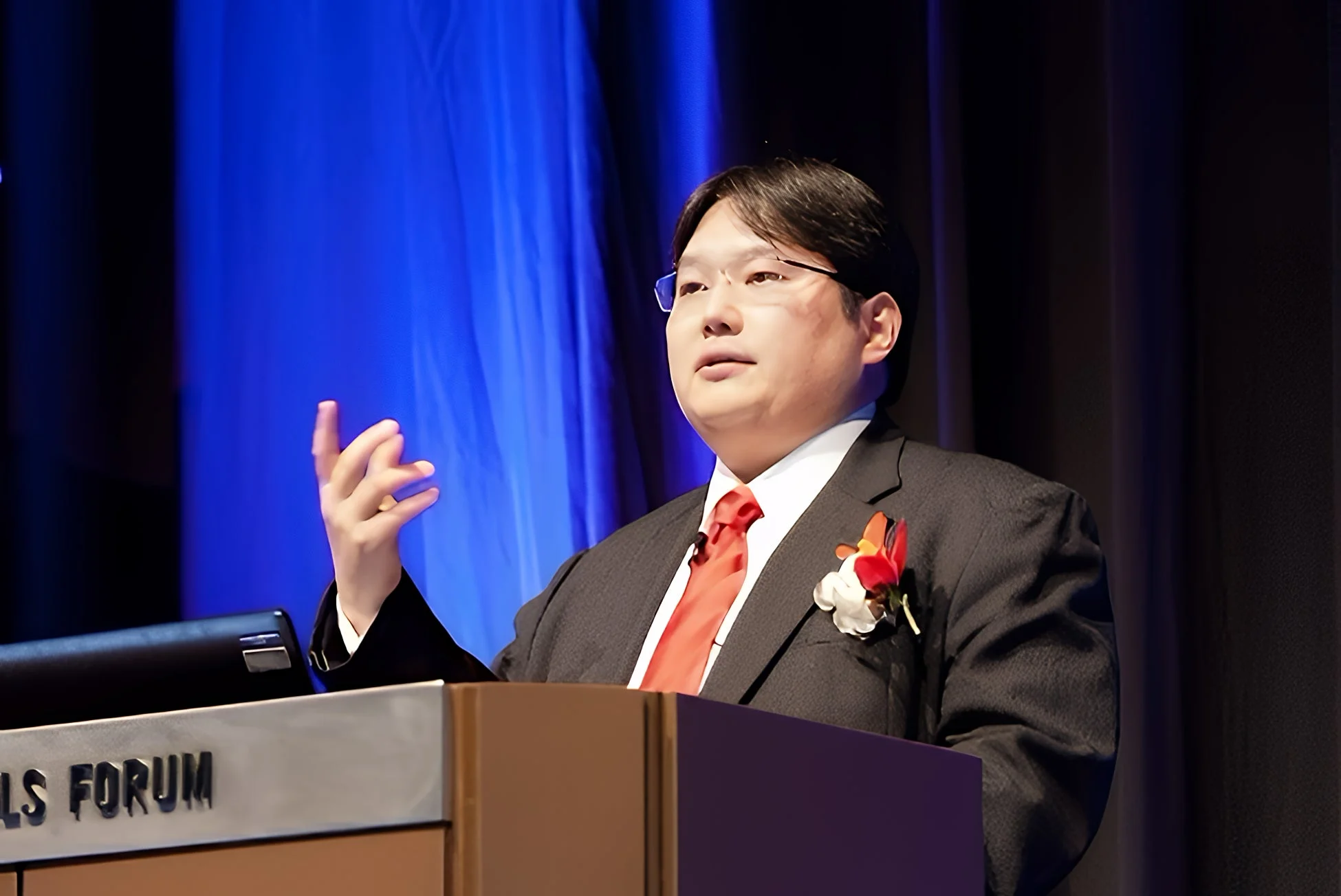 スーツを着て眼鏡をかけた男性が、演壇で身振り手振りを交えながらスピーチをしている様子です。背景は青と黒のカーテンで、フォーラムのようなイベントであることがうかがえます。
