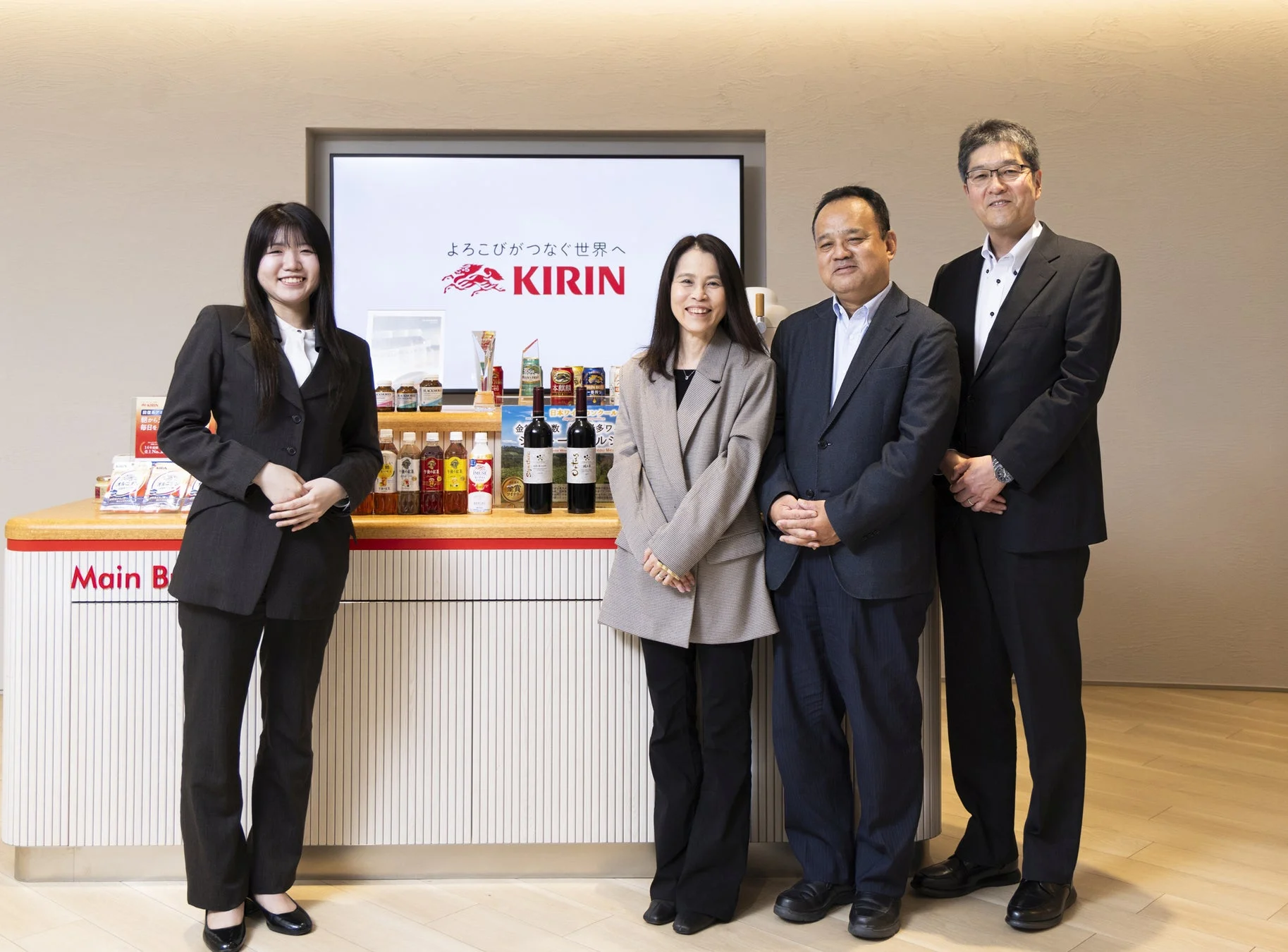 キリンのロゴと商品が並ぶカウンターの前で、4人のビジネスパーソンが笑顔で立っている集合写真。