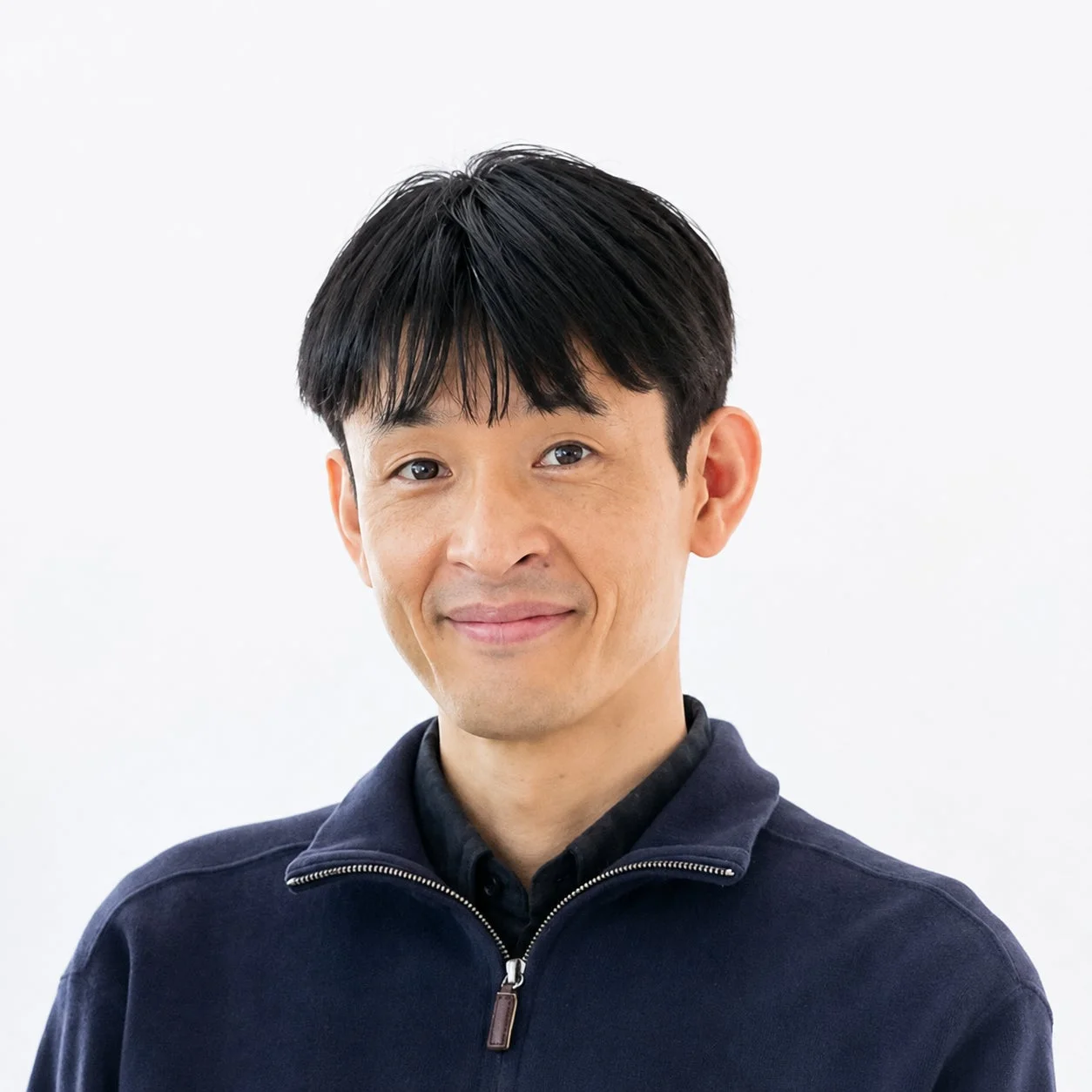 白い背景の前で、紺色のジップアップジャケットを着用したアジア系の男性が、穏やかな笑顔を浮かべている上半身のポートレートです。