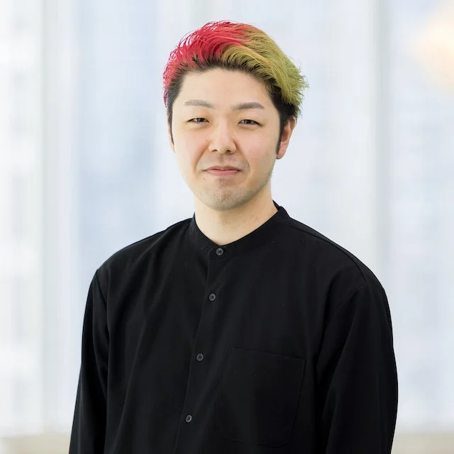 カラフルな赤と緑の髪が特徴的なアジア人男性のポートレート。黒い襟付きシャツを着用し、カメラをまっすぐ見つめています。背景は明るくぼかされた屋内空間です。